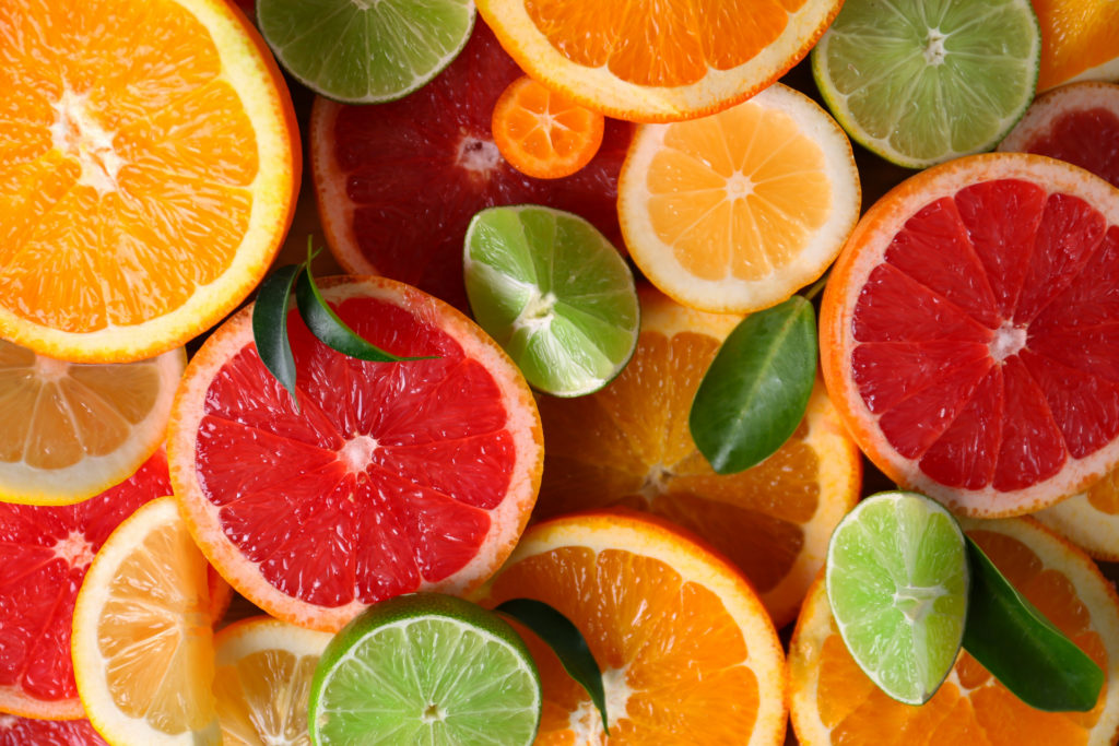 Various sliced citrus fruits including oranges, grapefruits, limes, and lemons, are arranged closely together with green leaves scattered throughout.