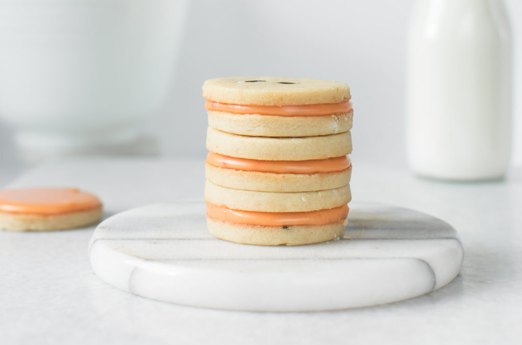 A stack of sandwich cookies with creamy orange filling sits on a white marble platter. A bottle of milk is blurred in the background, and an additional cookie is partially visible to the side.