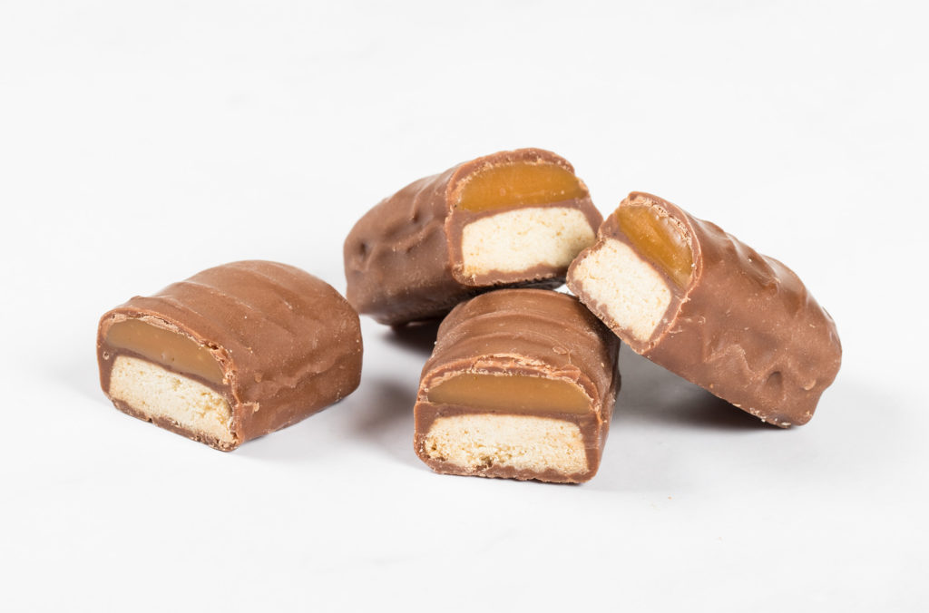 Four pieces of chocolate bars are on display, two of which are halved to reveal a layered filling of caramel and a biscuit base. The glossy surface hints at a smooth, creamy texture.