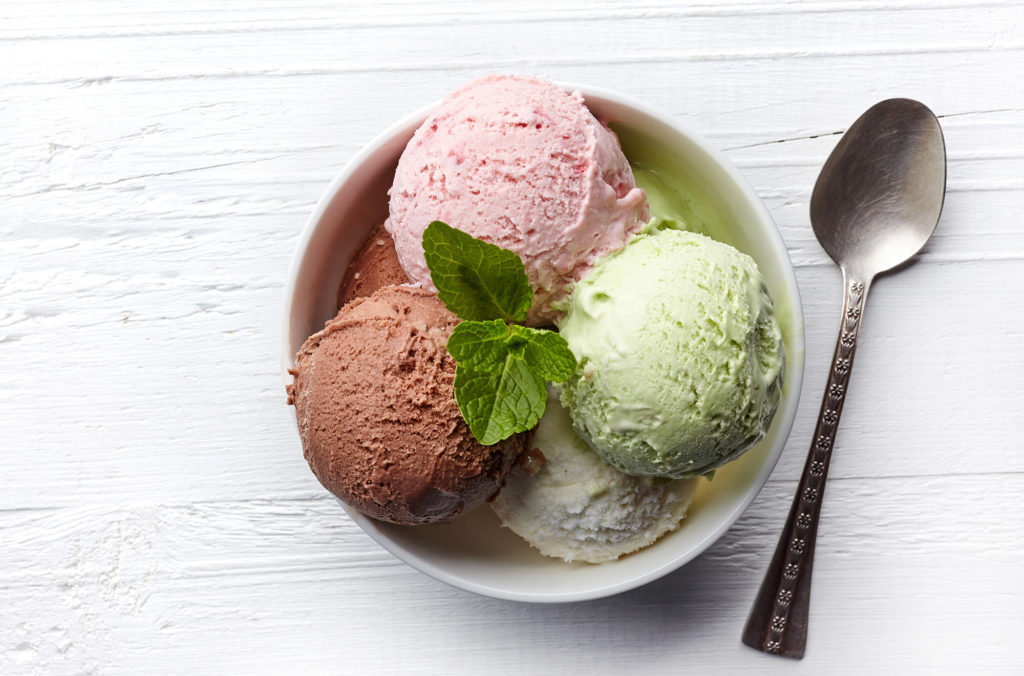 A bowl of ice cream with four scoops: pink strawberry, green mint, white vanilla, and brown chocolate. Garnished with a sprig of mint. A silver spoon lies beside the bowl on a white wooden surface.