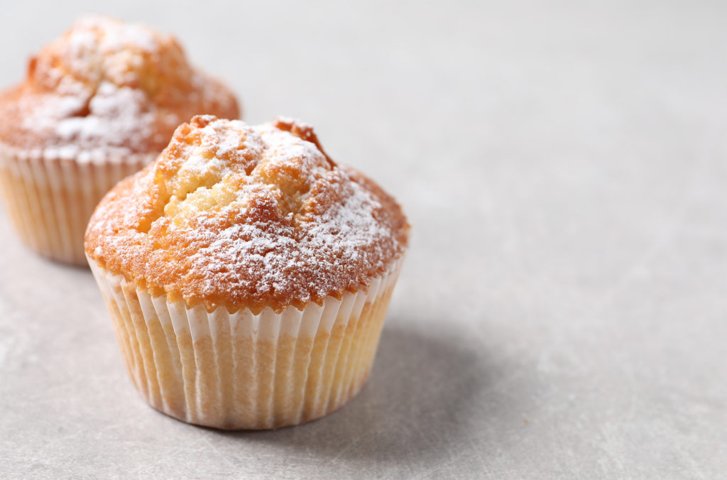 Two freshly baked muffins dusted with powdered sugar are placed on a textured gray surface. The muffins have a golden-brown top, and the background is softly blurred, highlighting the muffins' details.