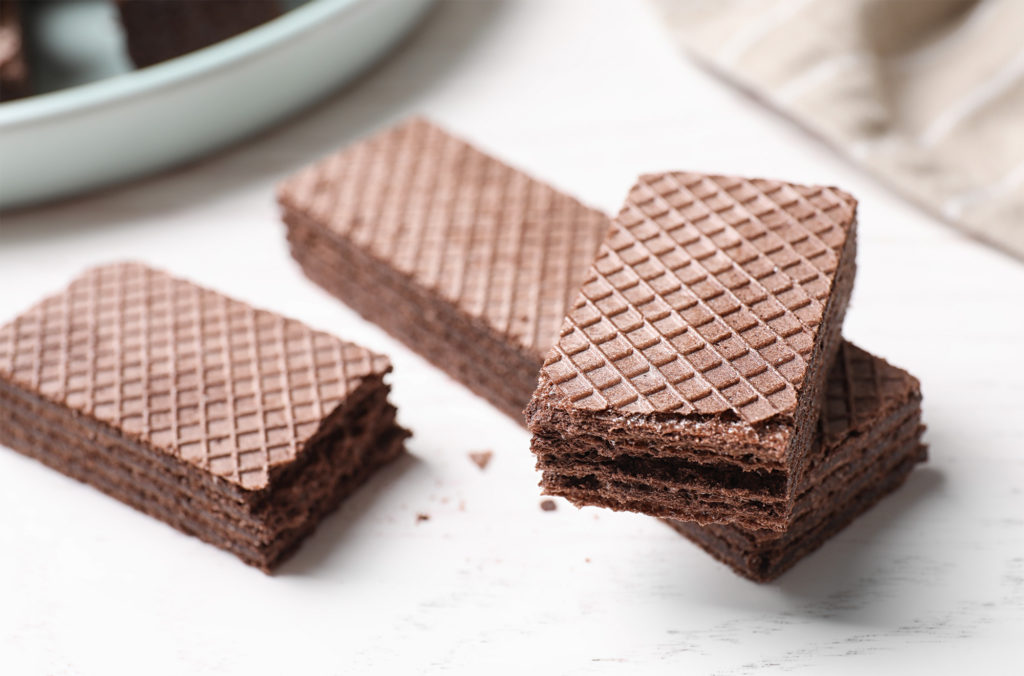 Three chocolate wafers are stacked on a white surface. The wafers have a textured, diamond-patterned top. One wafer is bitten, revealing layers of chocolate filling. A blurred piece of cloth is visible in the background.