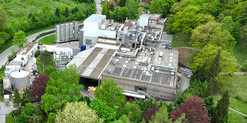 Aerial view of an industrial building surrounded by lush green trees and vegetation. The facility has large rooftops, several vents, and is situated near a winding road.