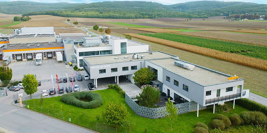 Aerial view of a modern industrial complex surrounded by green fields and farmland. The complex consists of two white buildings connected by a covered walkway. Several cars and trucks are parked outside. Forested hills are visible in the background.