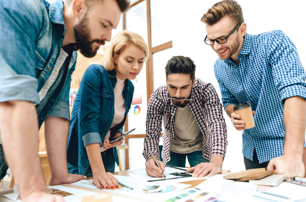 A group of four people gather around a table, collaborating on a project. They are focused on documents and design materials, with one person writing. Casual attire suggests a creative or informal workplace.