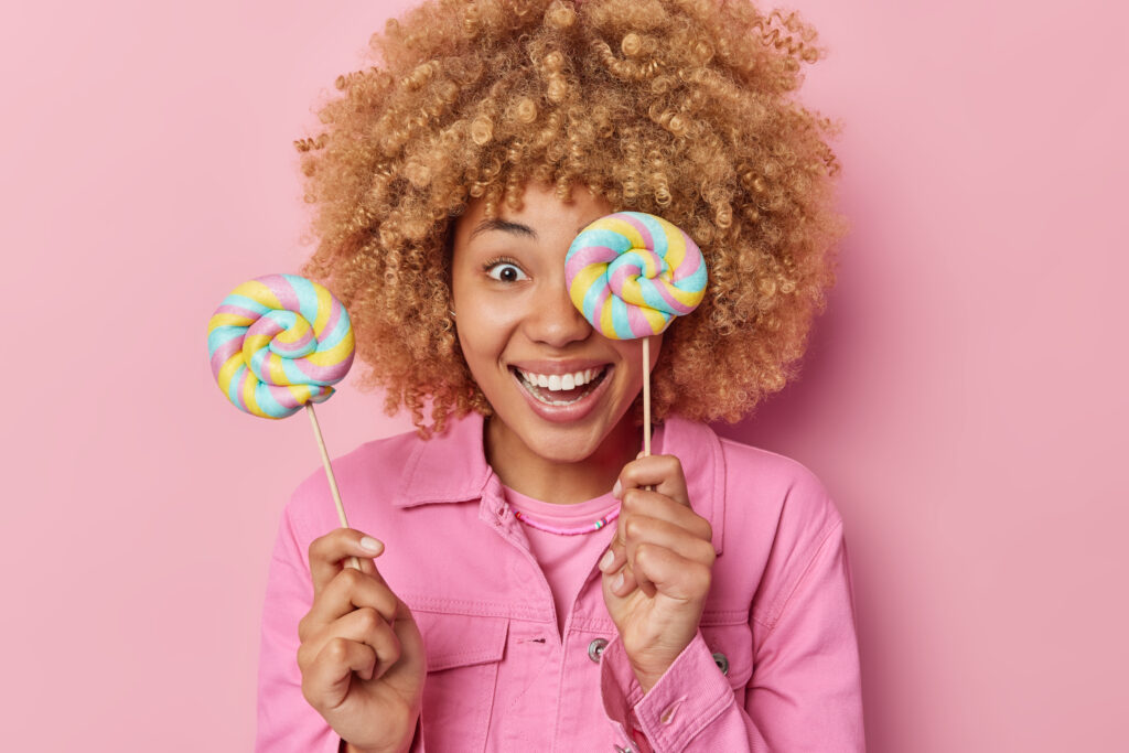 Smiling glad woman with curly hair holds sweet lollipops on sticks enjoys eating favorite treats wears pink jacket poses indoor. Positive female model eats caramel candies rewards herself with sweets